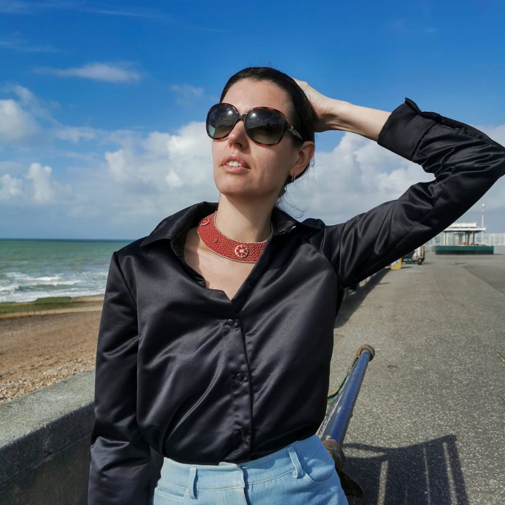 Woman wearing a red and gold boho collar necklace with medieval heritage design, styled with a black silk shirt and light blue trousers by the seaside.