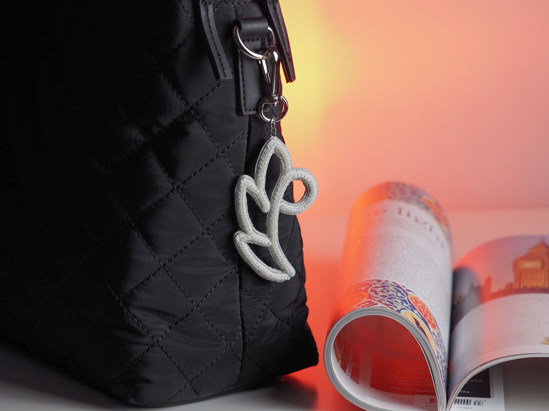 Handmade white and silver floral bag charm with seed beads and metallic cord, shown attached to a black quilted handbag
