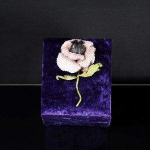 One-of-a-kind blush pink poppy brooch made of sequins and embroidered threads, displayed upright on a purple velvet stand