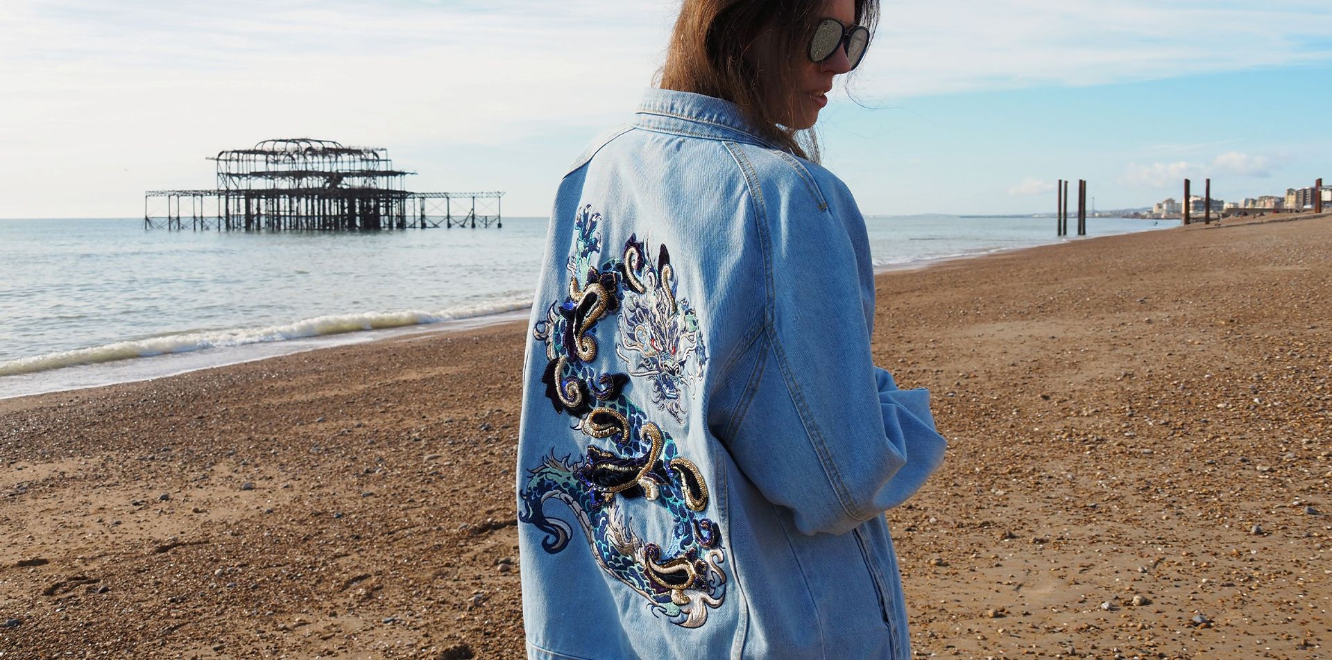 Woman wearing an oversized denim jacket with hand-embroidered dragon motif, standing on Brighton beach near the West Pier. Banner image for embroidered dragon denim jacket product by BY ROVA
