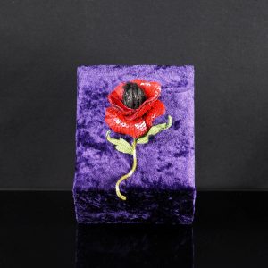 One-of-a-kind red poppy textile brooch with sequins and embroidered stem, displayed upright on a purple velvet stand