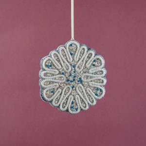 Hanging view of the Jewels of Winter circular snowflake ornament by Ksenia Semirova, crafted with pearl-setting technique in white, silver, and blue tones. Shown suspended on a satin ribbon against a pink background.