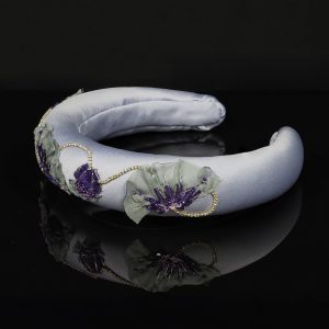 Side view of a lavender satin padded headband with hand-stitched poppy motifs in purple sequins and grey chiffon, decorated with rhinestone chain, resting on a black reflective surface
