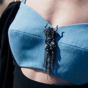 Taurus textile brooch with black embroidery and chain fringe, worn on a blue structured top as a statement accessory